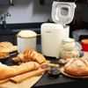 White bread maker with open lid on kitchen counter, surrounded by fresh bread and baking ingredients