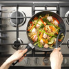 Stir fry with salmon, noodles and vegetables in frying pan on gas hob, hands holding utensils