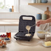 Black waffle maker open, batter being poured in, kitchen ingredients visible.