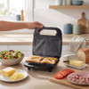 Black sandwich toaster making two toasted sandwiches with salad and toppings nearby