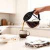 Glass coffee carafe with black handle pouring coffee into mug on kitchen counter