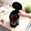 Black filter coffee machine with open lid, ground coffee being added with scoop