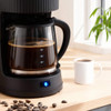 Black filter coffee machine with open lid, ground coffee being added with scoop