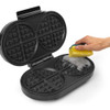 Open black waffle maker being cleaned with a yellow sponge