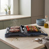 Open electric grill cooking four beef burgers in kitchen, ingredients prepared beside.