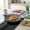 Blue frying pan with cooked salmon fillet, herbs and lemon slices on a modern hob.