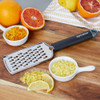 Stainless steel zester with black handle, shown with citrus fruits and grated zest.