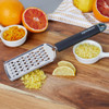 Stainless steel zester with black handle on wooden board, shown with citrus zest and sliced lemon.