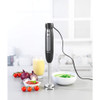 Hand blender mixing green soup in bowl, with tomatoes, onions, and juice on kitchen counter