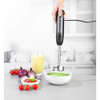 Hand blender mixing green soup in bowl, with tomatoes, onions, and juice on kitchen counter