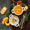 Two bowls of homemade soup with bread slices, fresh herbs, and vegetables on a rustic tray
