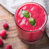 Glass of orange juice with two green leaves, served on a white plate with raspberries.