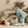 Teal kitchen mixer set with mixing bowl, tray, utensils, and baked goods on worktop