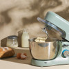 Teal stand mixer with whisk attachment and mixing bowl on worktop, baking ingredients nearby