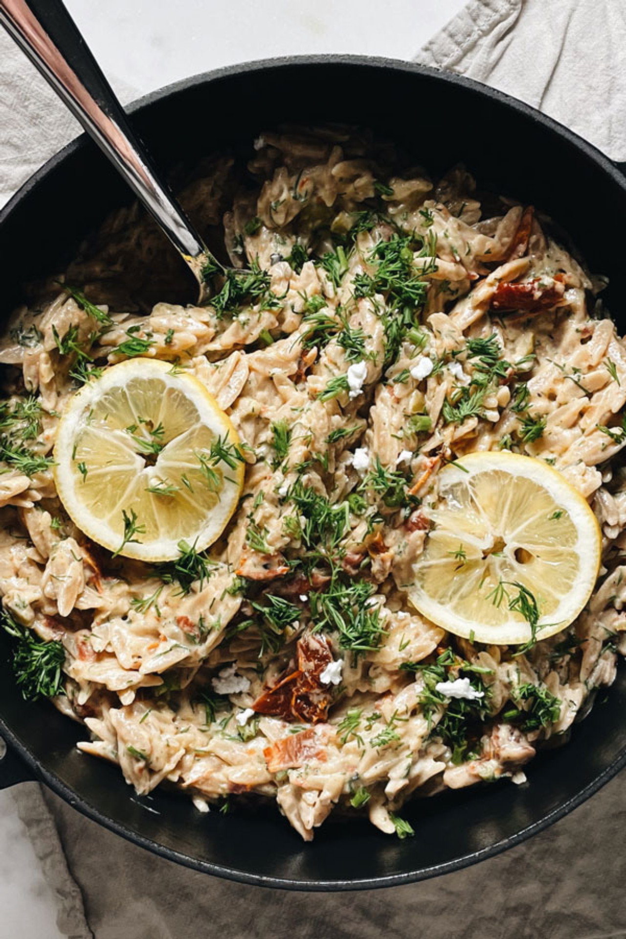 Creamy cheesy orzo pasta with sun-dried tomatoes, crumbled cheese, chopped olives, fresh dill and lemon slices served in a pot