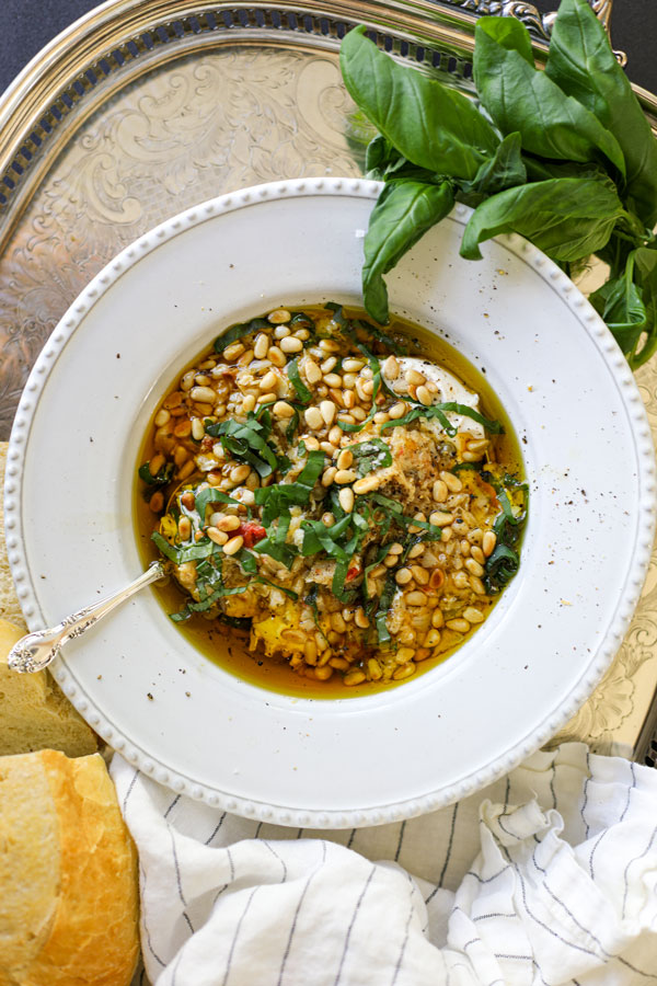 A white bowl filled with burrata artichoke bruschetta dip, garnished with fresh basil, pine nuts, and a drizzle of olive oil, served on a silver tray with fresh bread and basil on the side.