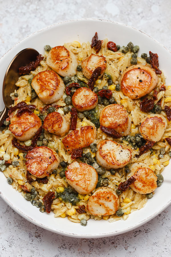 Orzo pasta tossed with sweet corn and sun-dried tomatoes topped with grilled scallops and served on a plate