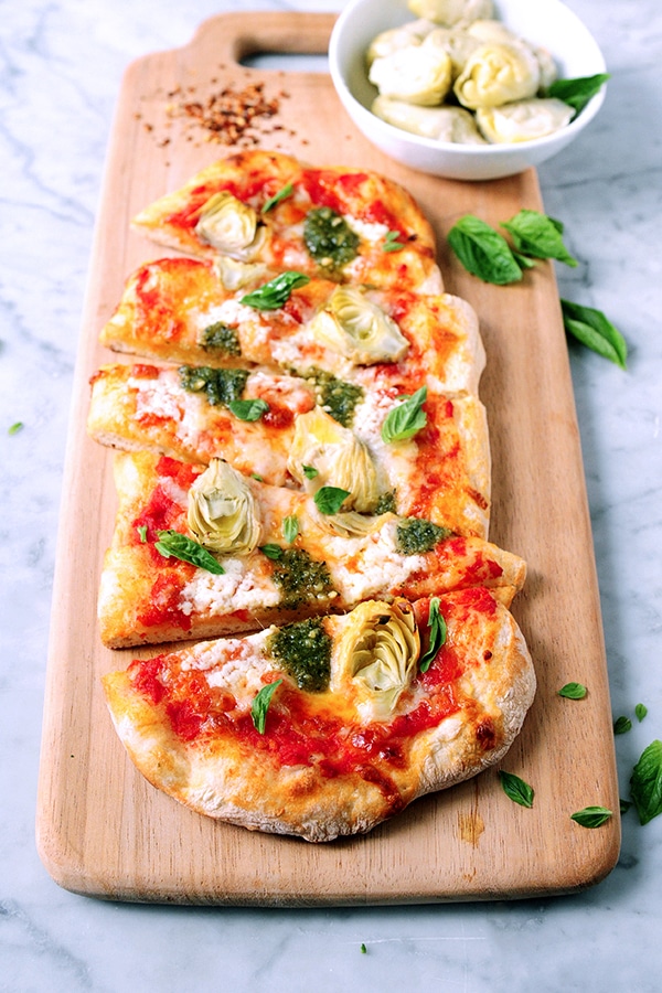 A pizza cut into slices served on a cutting board. The pizza is topped with artichokes and pesto. 