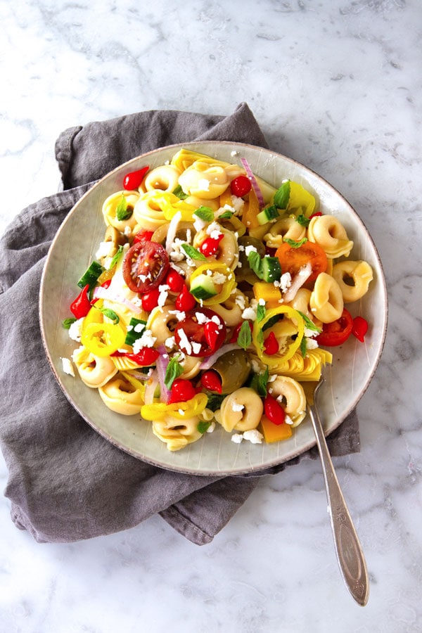 Tortellini pasta tossed in a bowl with tomatoes, cucumbers, olives and pepper drops