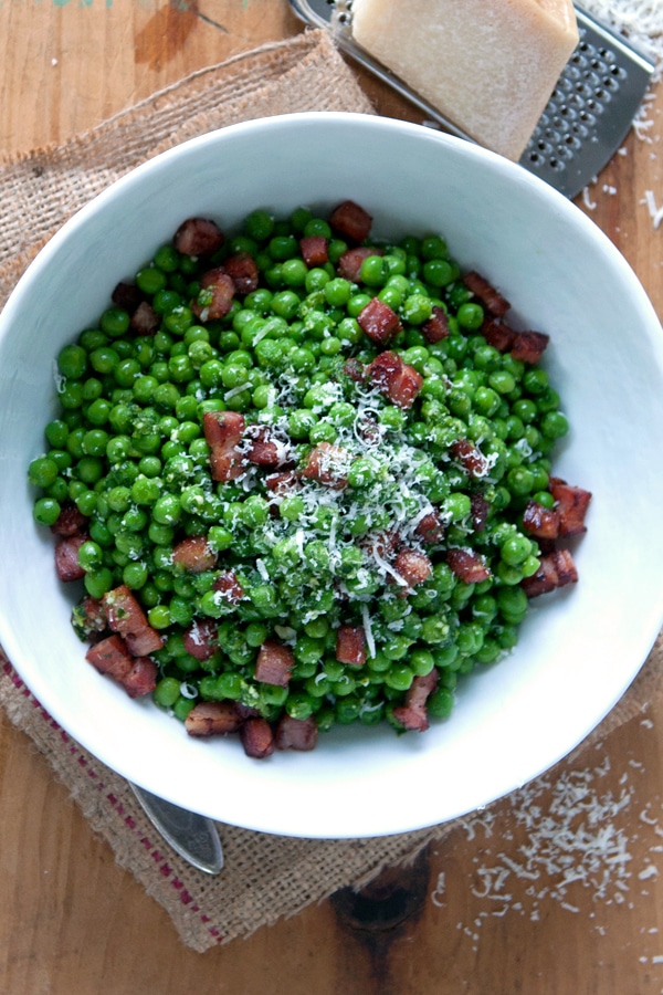 A bowl of peas with pancetta topped with some cheese. 