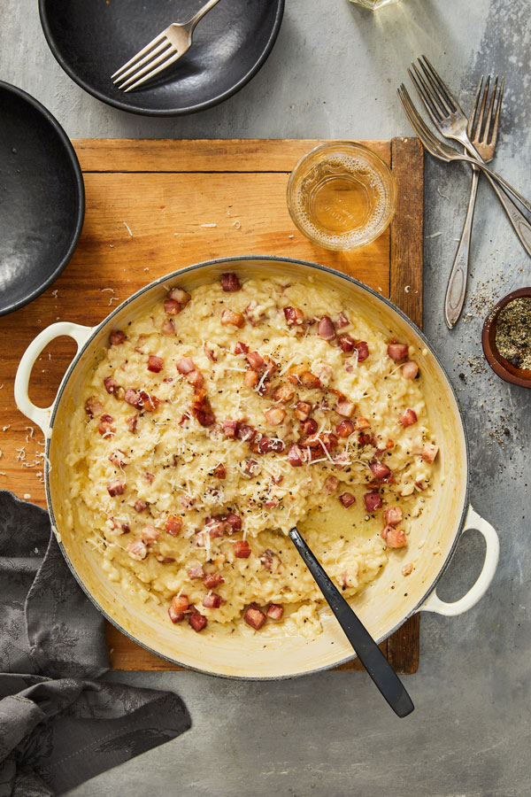 Cheesy risotto with diced pancetta and topped with grated cheese in a pot with a serving spoon.