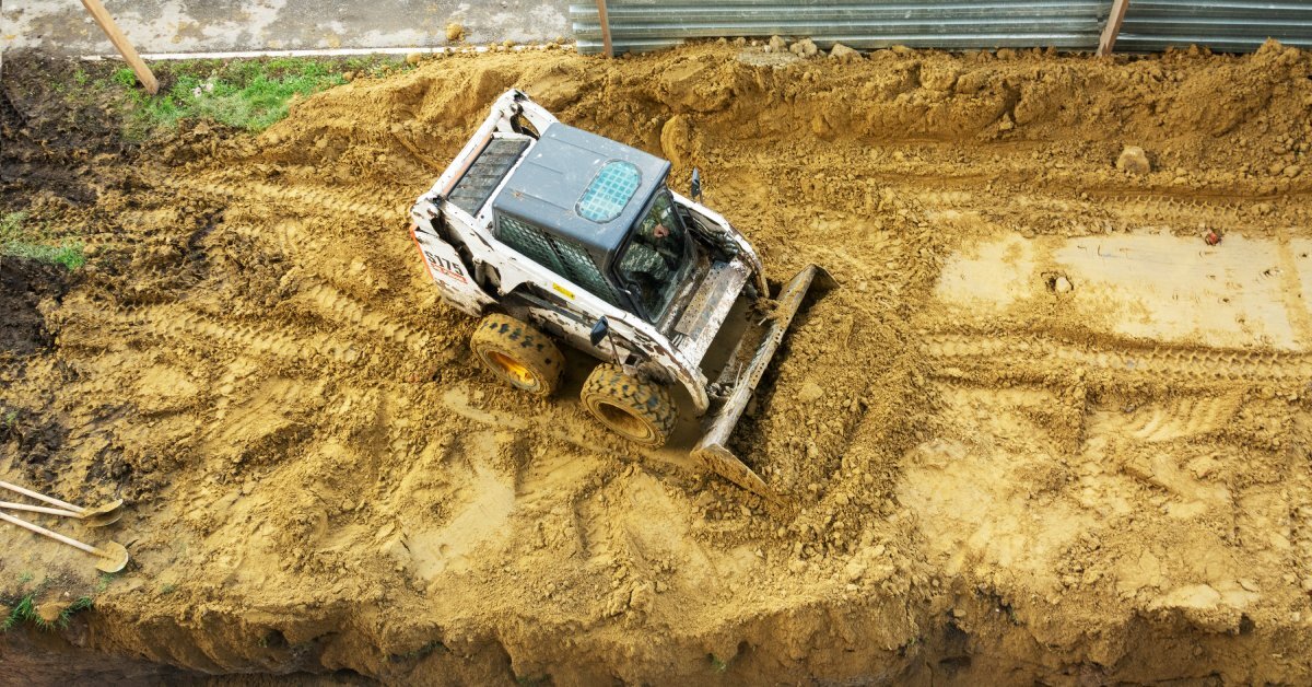 A compact track loader is in the middle of an unpaved outdoor area with imprinted tracks, loading up a pile of dirt.
