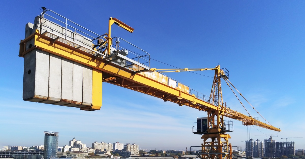 A massive yellow crane with two long arms has a large set of grey counterweights at the end of one end with a city skyline.