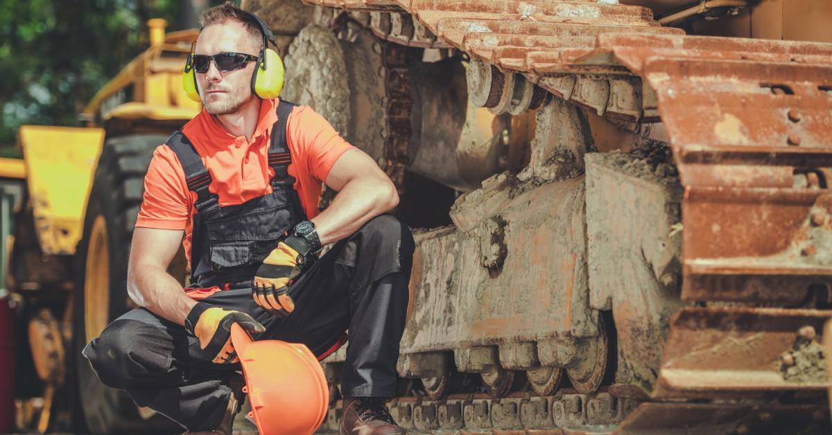 A repairman holding a hard hat and wearing ear muffs is crouched alongside the tracks of heavy machinery.