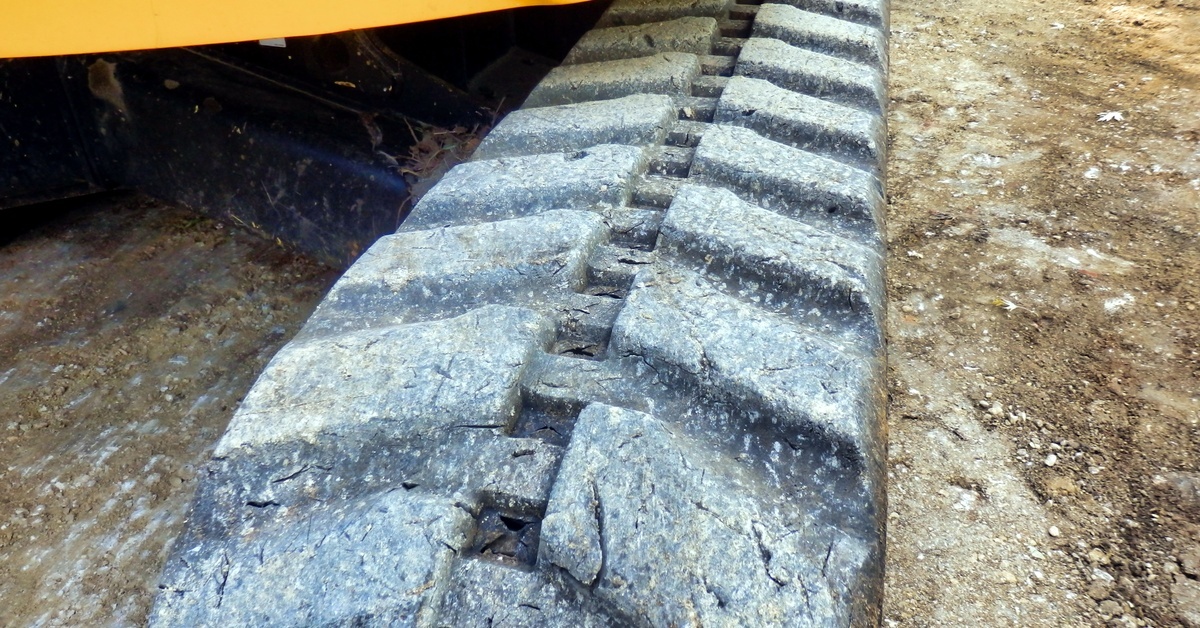 A close-up of cracked, dirty, worn-out rubber tracks on heavy machinery on dirt. A grassy field is in the distance.