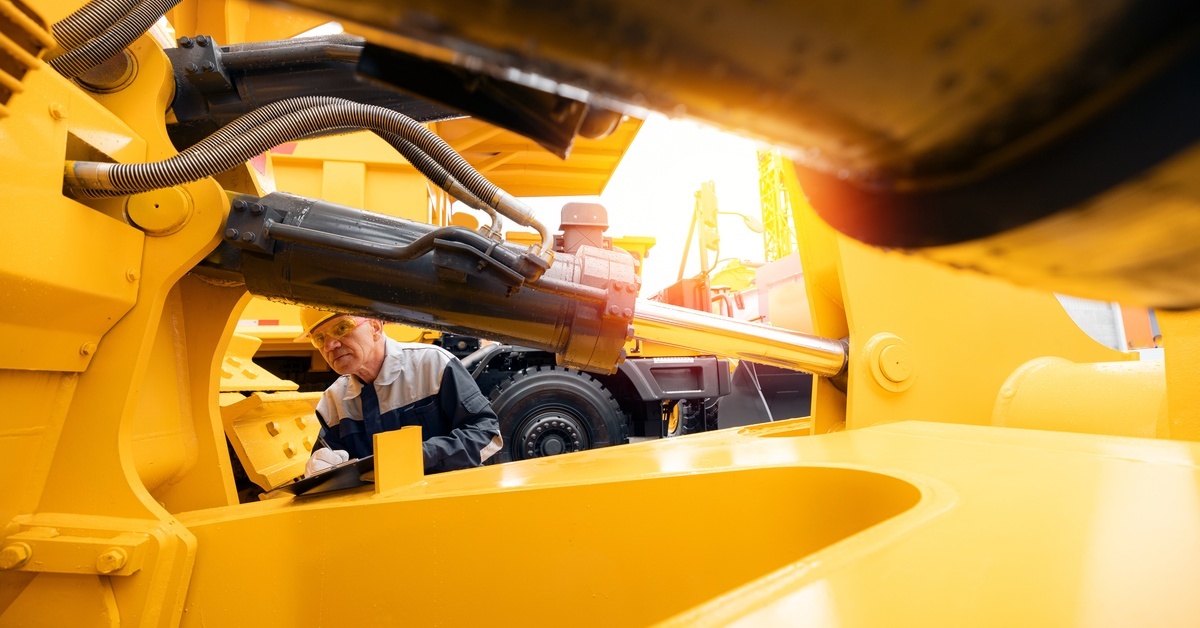 A close-up of heavy, yellow machinery, with a man wearing a uniform and holding a clipboard inspecting it nearby.