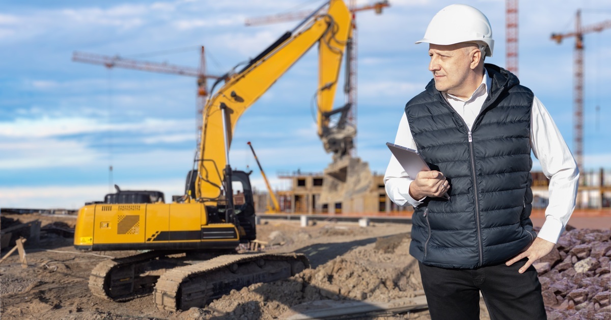 A man with a hard hat and a clipboard is looking over a construction site with heavy machinery and cranes behind him.