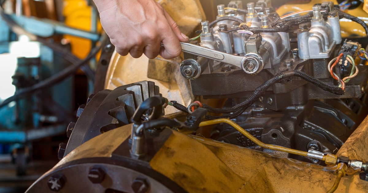 A person uses a wrench to tighten a component of a transmission within large yellow heavy machinery.