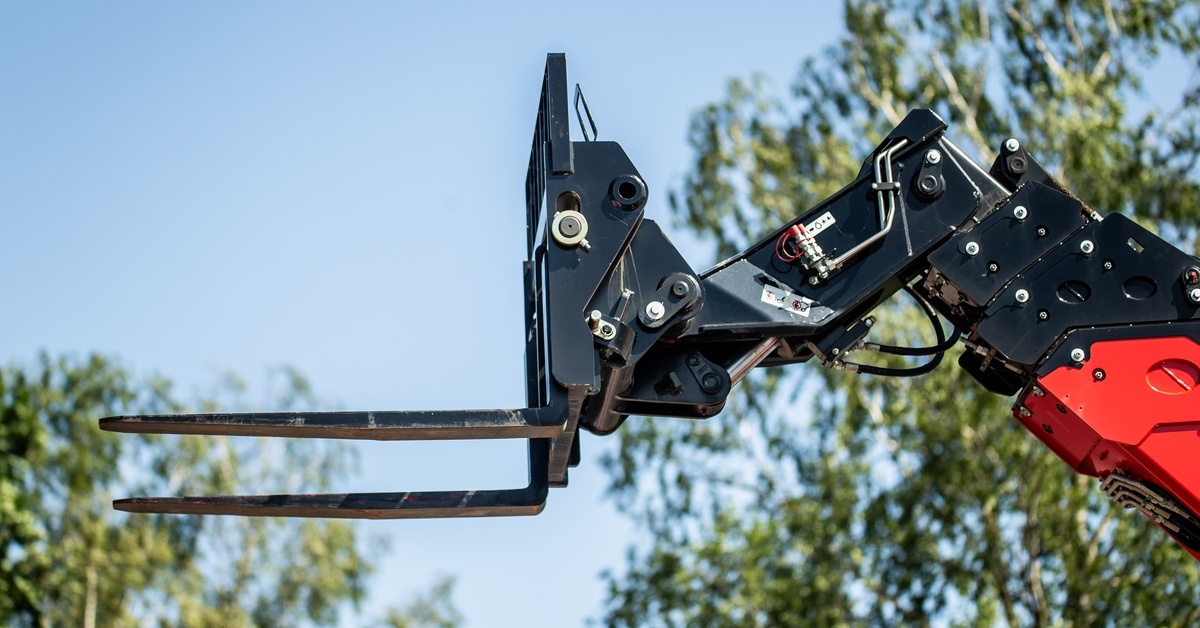 A close-up of a forklift attachment on the end of a red and black machine, with trees in the distance outside.