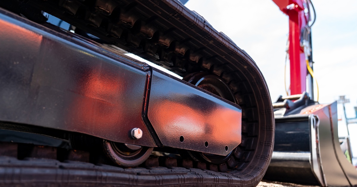 A close-up of the side of an excavator's rubber tracks sitting on gravel. An extension arm of the machine is nearby.