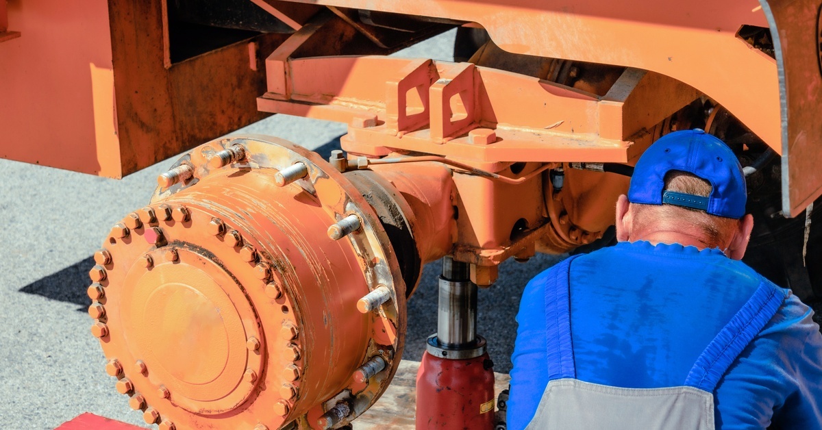 A close-up of an axle on a heavy machine, hoisted in the air. A worker wearing a uniform is alongside it.