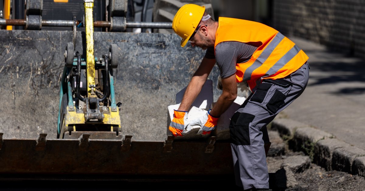 A construction worker bending forward and adjusting materials on the dirty bucket of an excavator on a city street.