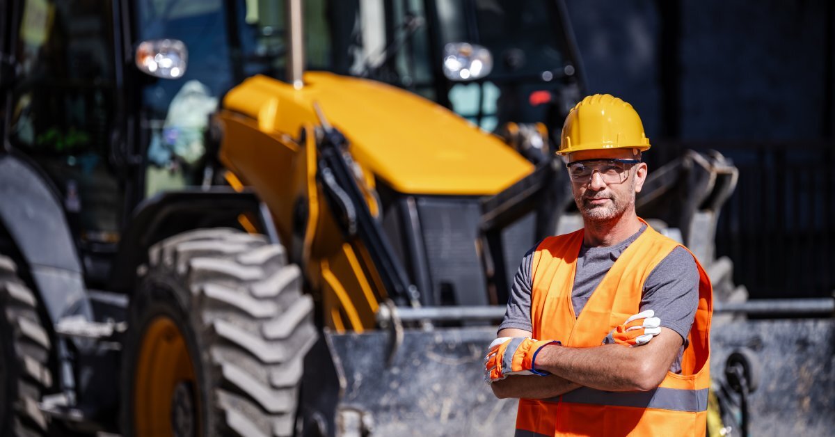 A construction worker standing with his arms crossed in front of an excavator. He is wearing a hard hat and orange vest.