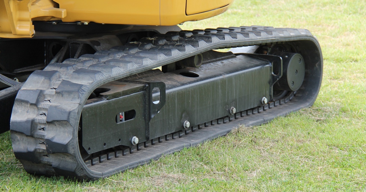 A close-up of rubber tracks on heavy yellow machinery. It is sitting on a grassy area. The tracks are dusty.