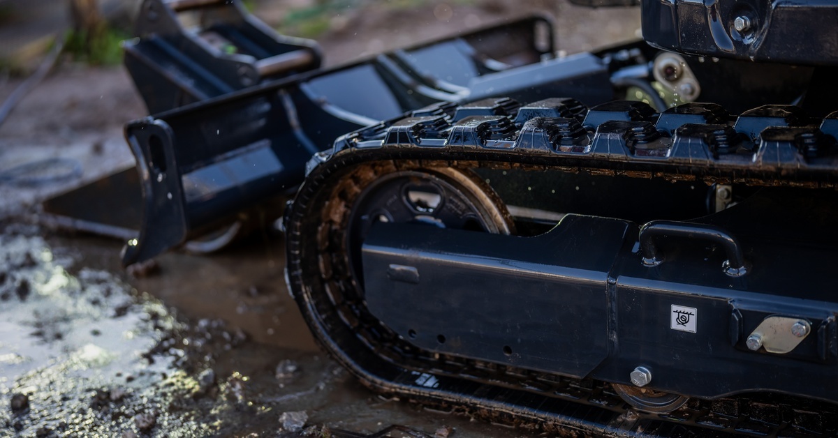A close-up of rubber tracks on heavy machinery. The tracks are treading on a watery, dirt ground. The machine has a bucket.