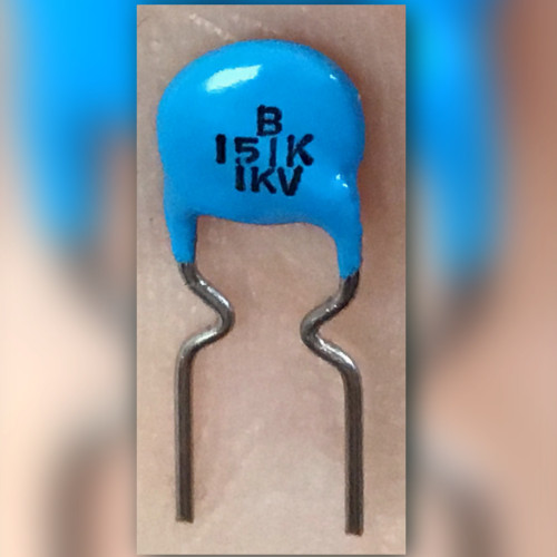 Blue 150pF 1KV ceramic capacitor marked B 151K held on a fingertip, front view