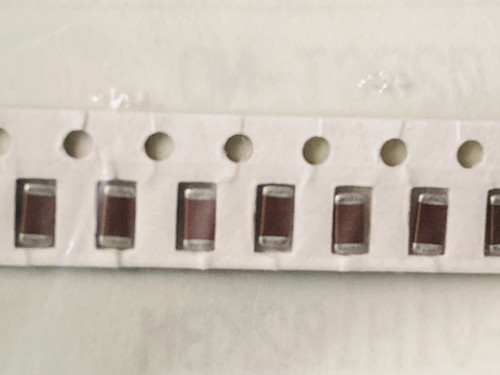 Row of brown ceramic chip capacitors held in white paper tape, sprocket holes above
