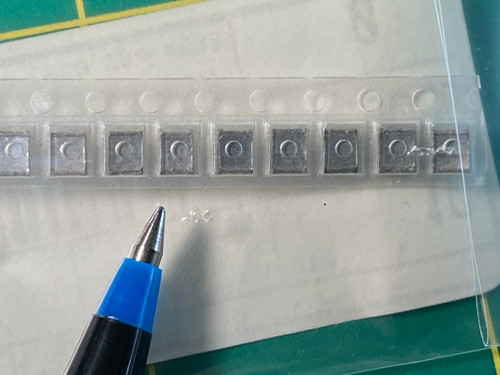 Nine squarish dark gray chip capacitors in clear carrier tape pockets beside blue pen