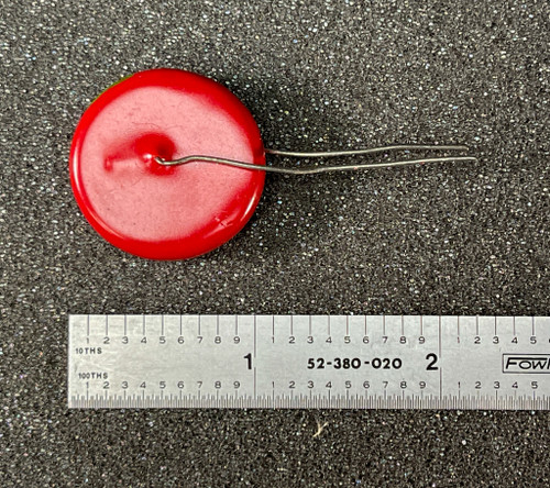 Mystery Ceramic Disc Capacitor 2000pF 10kV Radial Lead from PartsMine.com with a red circular body and two wire leads, placed next to a metal ruler for scale on a foam-like background.