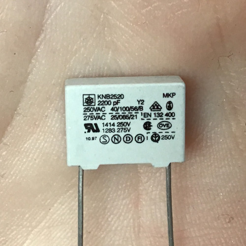 White rectangular correction capacitor (ISKRA KNB2520) with two metal leads, displaying printed specifications including "2200 pF," "250VAC," "275VAC," and various safety certifications like UL, VDE, and OVE.