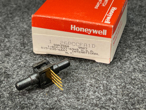 Close-up of a Honeywell Pressure Sensor 26PCCFA1D from PartsMine.com placed next to its original red and white Honeywell packaging on a gray surface. The sensor is a small, black component with protruding pins for connectivity.