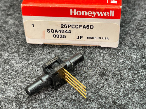 Honeywell Pressure Sensor 26PCCFA6D from PartsMine.com placed on a gray surface beside its red and white Honeywell packaging box, with the part number "26PCCFA6D" and other specifications visible on the label. The sensor is a compact, black component with protruding golden pins.