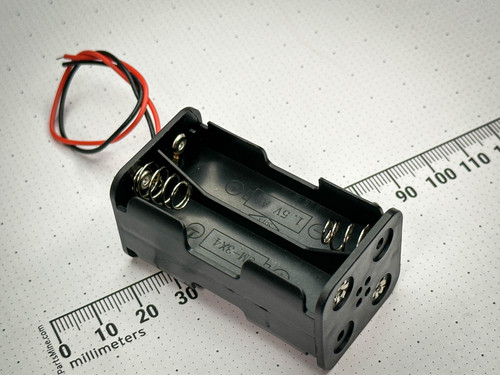 A 4 AA battery holder with black plastic casing and metal spring terminals, accompanied by red and black leads for connections. The holder is positioned beside a ruler marked in millimeters. Available at PartsMine.com.