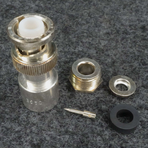 Disassembled connector showing main body with knurled coupling ring and white dielectric insert, threaded bushing, center pin, washer, and rubber grommet. Stamped code visible on body.