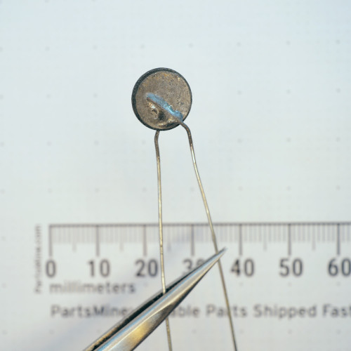 Bare disc PTC thermistor held with tweezers above a metric scale.