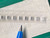 Row of tiny silver-gray chip capacitors seated in clear carrier tape, pen tip below